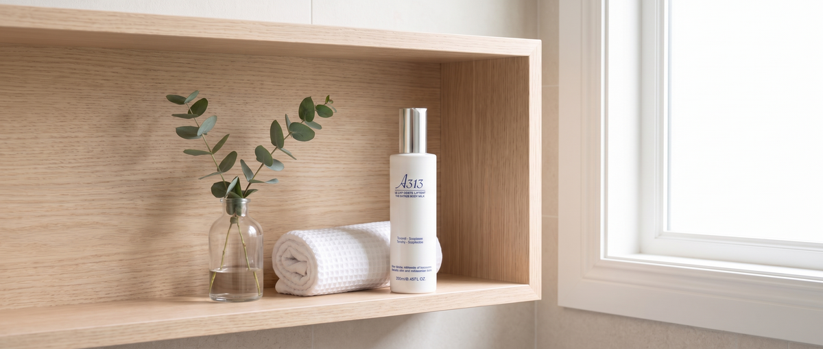 Flasche A313 Lifting Body Milk auf einem hellen Holzregal im Badezimmer neben einem gerollten weißen Handtuch und einem Eukalyptuszweig